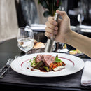 A hand uses the DDR BBQ Supply Thumb Grinder by DDR BBQ Supply to season a gourmet meat and veggie dish on a white plate, with a glass of water, knife, fork, and elegant table setting for a refined dining experience.
