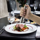 A hand seasons gourmet sliced meat, vegetables, and sauce with a DDR BBQ Supply Thumb Grinder—an essential one-handed tool by DDR BBQ Supply. Nearby on a black placemat rest a glass of water, fork, and knife.