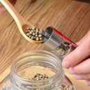 A hand uses the DDR BBQ Supply Thumb Grinder above a jar for easy one-handed seasoning, while a wooden spoon with peppercorns nears—ideal DDR BBQ Supply accessory for your BBQ on a wooden surface.