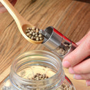 A hand fills a DDR BBQ Supply Thumb Grinder with whole peppercorns over an open glass jar, using a wooden spoon on a wooden surface.