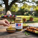 A hand dips a tortilla chip into a bowl of DDR BBQ Supply Tomatillo Salsa (Medium) Pint – 16 oz beside its jar. Guacamole, grilled skewers, and a colorful sombrero are arranged on an outdoor table in the sunshine.