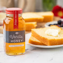 A jar of Honey Truck Co Raw Florida Vanilla Infused Honey rests next to slices of buttery pound cake on a white plate. Fresh berries and a red flower blur softly in the background.
