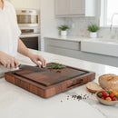 In a bright kitchen, someone slices fresh rosemary on the BBQ Boards® Best BBQ Board® 16 x 12 Cutting Board. Sliced bread, black peppercorns, cherry tomatoes, and olives sit on the marble countertop.