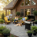 Four adults enjoy a barbecue on a log cabin patio; two are seated in Kanyon Living Adirondack Poly Glider Swivel Chairs in Yellow & Cedar, while two stand with plates. A golden retriever lies nearby among lush plants and Amish-made outdoor furniture.