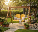 The Kanyon Living Poly Lumber 4′ Adirondack Swing in yellow and cedar wood features two chairs with cushions and a blanket on a stone patio, surrounded by potted plants, a small table with a teacup, lush greenery, and a house in the background.