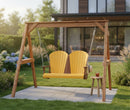 The Kanyon Living Poly Lumber 4′ Adirondack Swing in yellow and cedar wood hangs from a wooden frame in a garden, with a small stool holding iced tea nearby. The modern house and lush landscaping create an inviting outdoor setting.