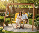 A smiling couple relaxes on the Kanyon Living Poly Lumber 4′ Adirondack Swing in yellow and cedar wood, enjoying a sunny backyard garden filled with flowers and greenery.