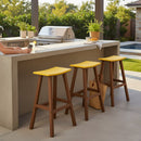 Outdoor kitchen with a concrete counter, weather-resistant furniture, three Kanyon Living Poly Lumber Saddle Stool Bar Height Yellow & Cedar Wood stools, grill, potted plants, pool in the background. Someone's arm holding a phone rests on the counter.