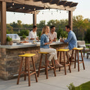 Four people chat and enjoy drinks at an outdoor bar and grill, relaxing on Kanyon Living Poly Lumber Saddle Stool Bar Height Yellow & Cedar Wood seating, surrounded by string lights, lush plants, and weather-resistant decor for a summery vibe.