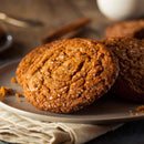 A close-up of three brown, crispy cookies topped with The Spice Lab’s Sanding Con AA Sugar, their sparkling crystals visible as they rest on a beige plate atop a folded napkin, with a blurred glass of milk in the background—a delicious showcase of decorative baking.