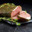 A slab of herb-crusted cured meat with a slice cut, garnished with The Spice Lab Organic Whole Thyme, sits on a black slate platter against a dark background—perfect for inspiring French, Italian, and Mediterranean cooking and pairing beautifully with your favorite seasoning blends from The Spice Lab.