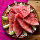Slices of watermelon sprinkled with The Spice Lab Ground Cayenne Pepper are arranged on a decorative plate, surrounded by lime wedges. The plate is set on a wooden table next to a pink striped cloth.