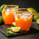 Two orange cocktails with ice, rimmed with The Spice Lab Chili Lime Margarita Salt Cocktail Rimmer by The Spice Lab, and garnished with lime wedges and jalapeño slices, sit on a slate surface alongside extra lime slices and jalapeño pieces.