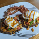 Two slices of seeded toast topped with diced tomatoes, onions, bell peppers, poached eggs, and Terrapin Ridge Farms Spicy Chipotle Garnishing Squeeze, served with crispy bacon strips on a white plate.