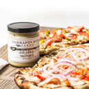 A jar of Terrapin Ridge Farms Dill Pickle Mustard sits on a wooden board beside two flatbread pizzas topped with vegetables like red onions and tomatoes.