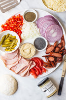 A plate with pizza toppings—sliced red onions, chopped red bell peppers, diced onions, tomatoes, ham, sausages, pickled peppers, poppy seeds, and ground black pepper—with Terrapin Ridge Farms’ Dill Pickle Mustard and pizza dough nearby.