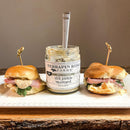 Two ham and cheese slider sandwiches with lettuce flank an open jar of Terrapin Ridge Farms Dill Pickle Mustard on a white rectangular plate, with a spoon resting inside the savory condiment jar.