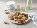 A plate of crostini with prosciutto, cheese, fig jam, and herbs sits on a table with two glasses of white wine and a jar of Terrapin Ridge Farms Balsamic Onion Jam—the perfect cheese board condiment—nearby.