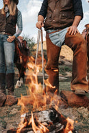 Two people in cowboy attire cook over a campfire using the Donkey Long Tong The Fire Master (32") by Donkey Long Tong | USA | CAN | AUS | UK, capturing classic Western outdoor cooking. Horses can be seen in the background, enhancing the rustic setting.