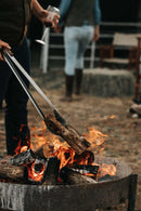 One person uses the Donkey Long Tong The Fire Master (32") from Donkey Long Tong | USA | CAN | AUS | UK to adjust firewood in a blazing outdoor fire pit, while another stands nearby holding a wine glass.