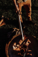 Wearing tan pants, a person uses the Donkey Long Tong The Fire Master (32”) by Donkey Long Tong to add wood to a blazing fire pit, with logs scattered on the grass—ideal for memorable campfire cooking.