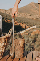 A hand holds the Donkey Long Tong The Fire Master (32") by Donkey Long Tong | USA | CAN | AUS | UK beside a wicker basket and an axe in a tree stump on a wooden deck, with scenic mountains in the background.