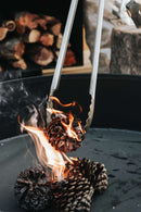 The Donkey Long Tong The Fire Master (32") from Donkey Long Tong | USA | CAN | AUS | UK easily holds a burning pine cone over a firepit with bright flames and smoke, while other pine cones and stacked firewood wait for campfire cooking.