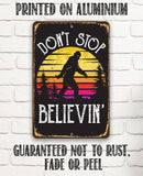 The Lone Star Art "Don't Stop Believing Big Foot" metal sign features a Bigfoot silhouette at sunset with trees. Text: DONT STOP BELIEVIN. PRINTED ON ALUMINUM. GUARANTEED NOT TO RUST, FADE OR PEEL—perfect for cabin decor.