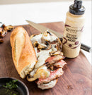 A steak sandwich with melted cheese and sautéed mushrooms on a wooden board, served with Terrapin Ridge Farms Bacon Aioli Quart for the perfect smoky spread, by Terrapin Ridge Farms.