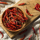 A wooden bowl brimming with Rico Rico Chile de Árbol Dried Chiles – Hot & Authentic Mexican Peppers rests on a wooden cutting board, while more of these fiery chiles from Rico Rico are scattered across a beige cloth for an authentic Mexican touch.