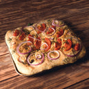 A rectangular focaccia bread topped with sliced tomatoes, red onion rings, and a sprinkle of The Spice Lab Ground Rosemary sits on a wooden surface.