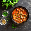 A skillet filled with chunky meat stew garnished with herbs sits surrounded by small bowls of chopped herbs and spices—including The Spice Lab Birdseye Chili Pepper Powder—along with some fresh parsley, all arranged on a dark surface.