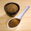 A black bowl filled with whole cumin seeds sits beside a white ceramic spoon containing The Spice Lab Ground Cumin 1 lb Bulk Foodservice Spice, both essential as foodservice spices, arranged neatly on a light wooden surface.