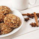 A plate of sesame and seed cookies sits on a white table next to a white mug, with cinnamon sticks and The Spice Lab Ground Star Anise—an aromatic spice from The Spice Lab known for its licorice-like flavor—scattered nearby.