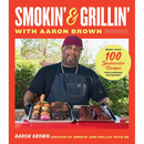 The cover of Smokin’ and Grillin’ with Aaron Brown: More Than 100 Spectacular Recipes for Cooking Outdoors features a man in a black apron holding two metal trays of grilled meats and vegetables.