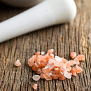 The Spice Lab Himalayan Pink Salt Coarse Grain by The Spice Lab is shown scattered across a rustic wooden surface, with a portion of a white mortar and pestle visible in the background—ideal for refilling your favorite salt grinder.