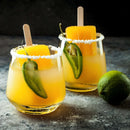 Two glasses with salted rims—each filled with vibrant yellow-orange drinks, popsicles, and sliced green chili peppers—are finished with a touch of The Spice Lab Chili Lime Margarita Salt Cocktail Rimmer by The Spice Lab and sit beside a fresh lime on a dark textured surface.