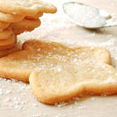 Butterfly-shaped sugar cookies topped with The Spice Lab’s Sanding Con AA Sugar are stacked and scattered on a light surface, with a spoon and more sparkling sugar in the background—a charming example of decorative baking.