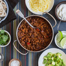 A pot of chili with beans sits on a striped cloth, surrounded by bowls of shredded cheese, sour cream, lime wedges, avocado, cilantro, salt, and flatbread. Seasoned with The Spice Lab Ground Cayenne Pepper for a touch of heat, the chili has a spoon resting inside.