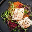 Two pieces of seasoned white fish rest on a bed of red sauce, garnished with assorted microgreens on a dark plate, their flavors enhanced by The Spice Lab Seafood Seasoning Set from The Spice Lab.