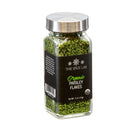 A transparent glass jar with a silver lid holds vibrant green The Spice Lab Organic Parsley Flakes, ideal for use as dried parsley or as a garnishing herb. Featuring a black label that displays “The Spice Lab Organic Parsley Flakes” and indicates a net weight of 0.5 oz (14g).