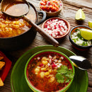 A bowl of red soup with hominy and meat, garnished with cilantro and chopped radish, sits on a plate next to a spoon, lime wedges, and small bowls of diced onion, radish, and salsa—all set for sprinkling with hot chile powder or The Spice Lab Chile de Arbol Powder for an extra kick—beautifully arranged on a wooden table.