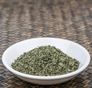 A white bowl filled with The Spice Lab Organic Sweet Basil—dried, chopped green herbs ideal for Italian-inspired dishes or as a savory poultry seasoning—rests on a woven brown surface.
