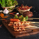 A wooden board displays assorted grilled skewers, sausage, and vegetables. In the background, bowls filled with dried chilies, spices, leafy greens, and a generous portion of The Spice Lab Minced Garlic 1 lb Bulk Foodservice Spice are set against a dark surface.