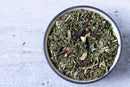 A bowl filled with dried green and brown herbs, including visible whole cloves, sits on a gray textured surface—perfect for the World Spice Loose-Leaf Tea Tasting Flight by World Spice or as part of an inviting herbal tea gift set.