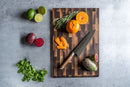 A Steelport Knife Co. Everyday Knife & Board Set featuring a wooden board and Made-in-USA kitchen knife, arranged with rosemary, orange peel, and an orange on top. Surrounding the set are limes, prickly pears, and mint displayed on a gray surface.