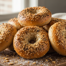 A pile of golden-brown bagels, topped with SALTOPIA Infused Sea Salts’ Everything Bagel Salt, rests on a wooden surface with scattered crumbs and soft natural light from a nearby window.