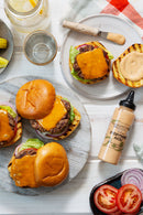 Overhead view of grilled gluten-free cheeseburgers with lettuce and cheddar on buns, sliced tomatoes, lemon water, and a bottle of Terrapin Ridge Farms Everything Aioli Squeeze Garnishing Sauce - 13 oz on a white table.