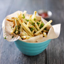 A turquoise bowl lined with parchment paper is brimming with thin, crispy French fries, topped with chopped herbs and a sprinkle of The Spice Lab Italian Black Truffle Sea Salt. The bowl rests on a wooden table, while a small dish of ketchup is softly blurred in the background.