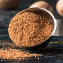 A metal measuring spoon filled with The Spice Lab Organic Ground Nutmeg, with some of the spice spilling onto a dark wooden surface—ideal for infusing warmth and depth into your eggnog or holiday baking.
